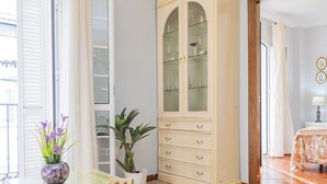 A room with a glass-top table, a wooden cabinet with glass doors, a potted plant, and a bed with a floral bedspread.