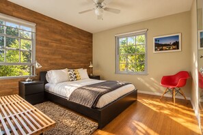 A bedroom with a bed, bedside tables, a desk, a chair, and large windows with a view of greenery.