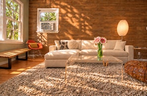 A modern living room with a sofa, coffee table, and a bench. There is a window with a view of trees, an air conditioning unit, and a lamp.