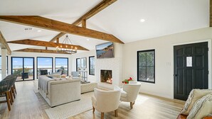 A spacious living room with a fireplace, large windows, and a high ceiling with exposed beams.