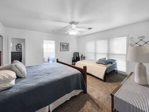 Une chambre avec deux lits, un ventilateur de plafond, une fenêtre avec des stores et une table de chevet avec une lampe.