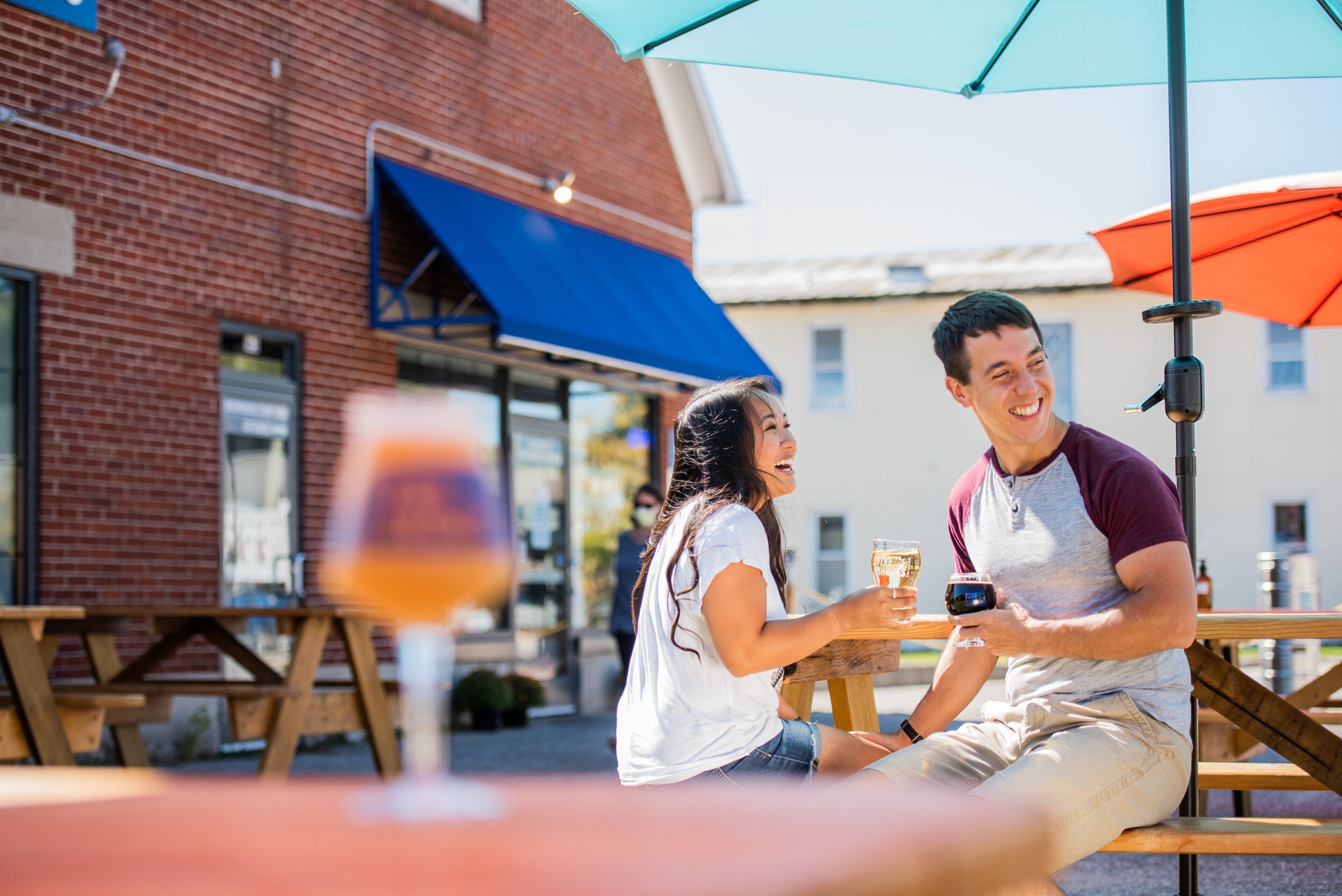 Two people sitting at an outdoor table with drinks, a brick building, and a blue awning in the background.