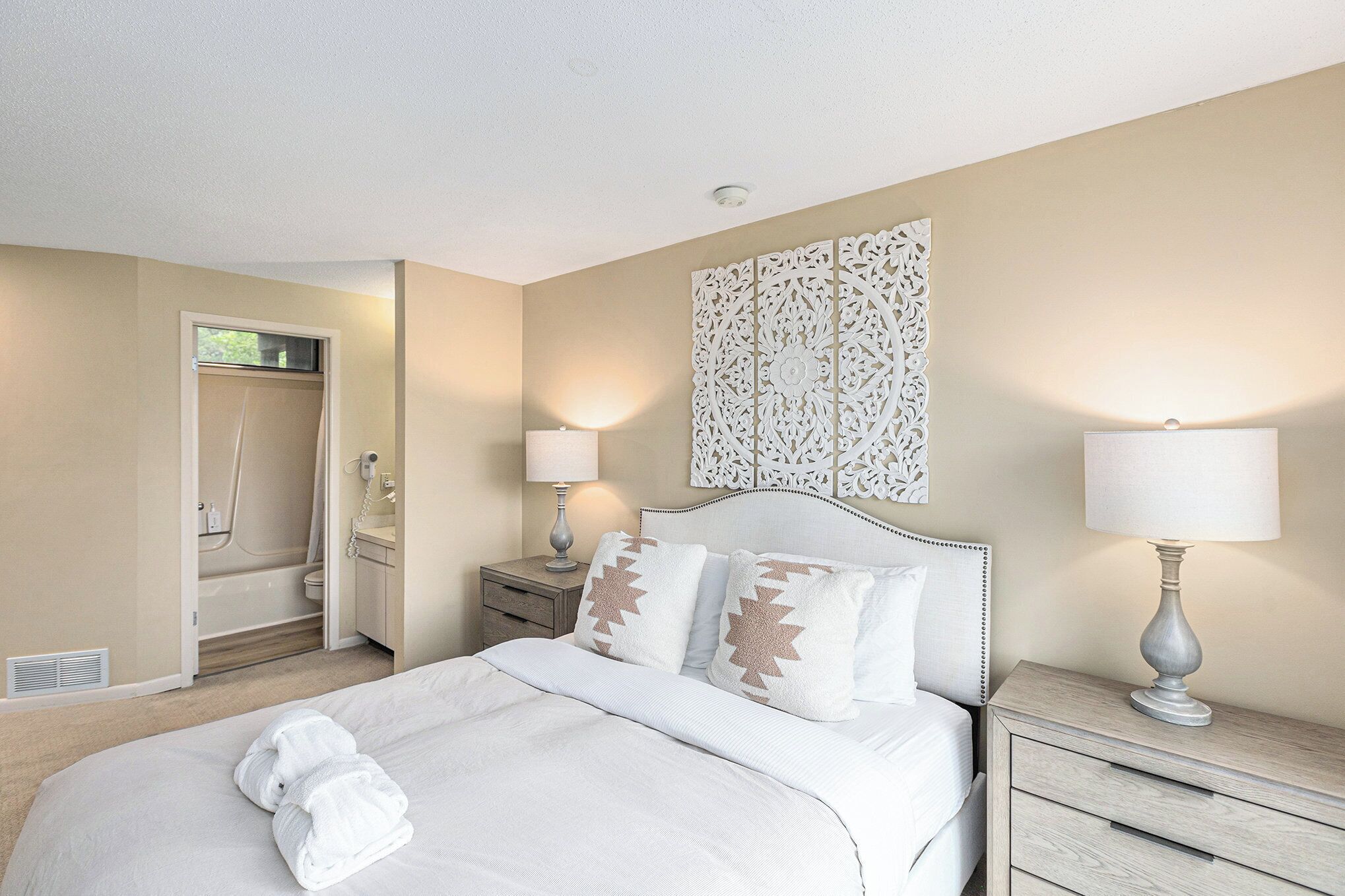 A hotel room with a bed, bedside lamps, a decorative wall piece, a bathroom with a shower, and a towel on the bed.