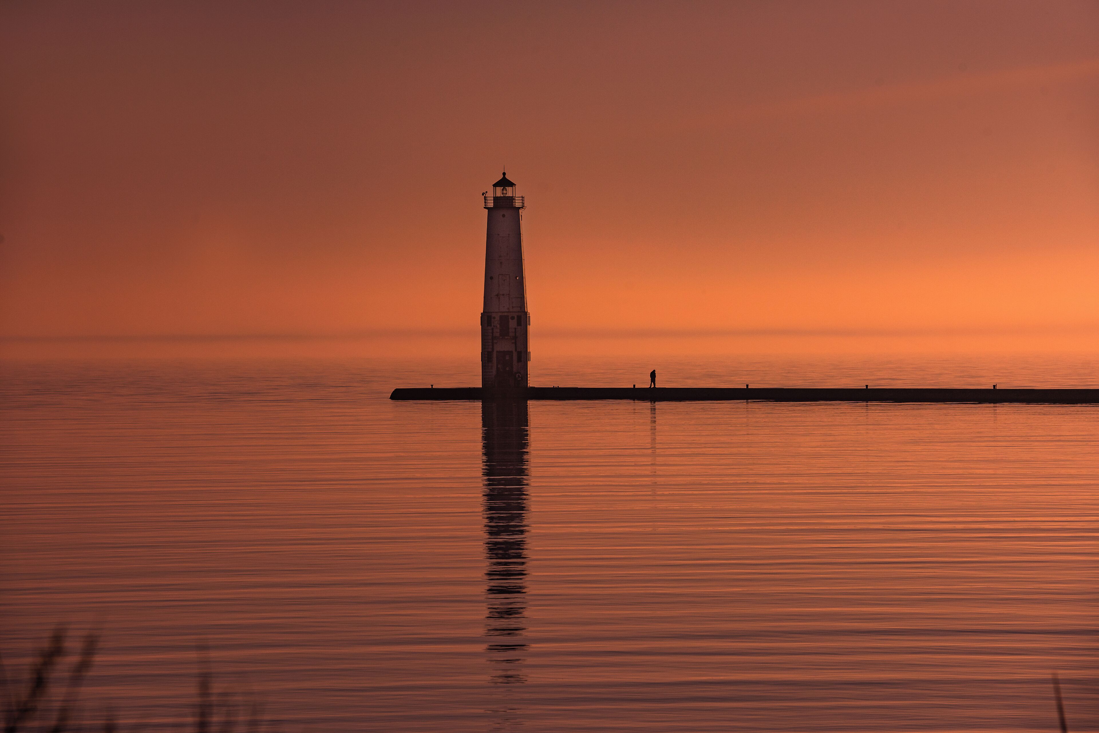 En silhuet af et fyrtårn mod en solnedgangshimmel, roligt vand og en fjer pier.