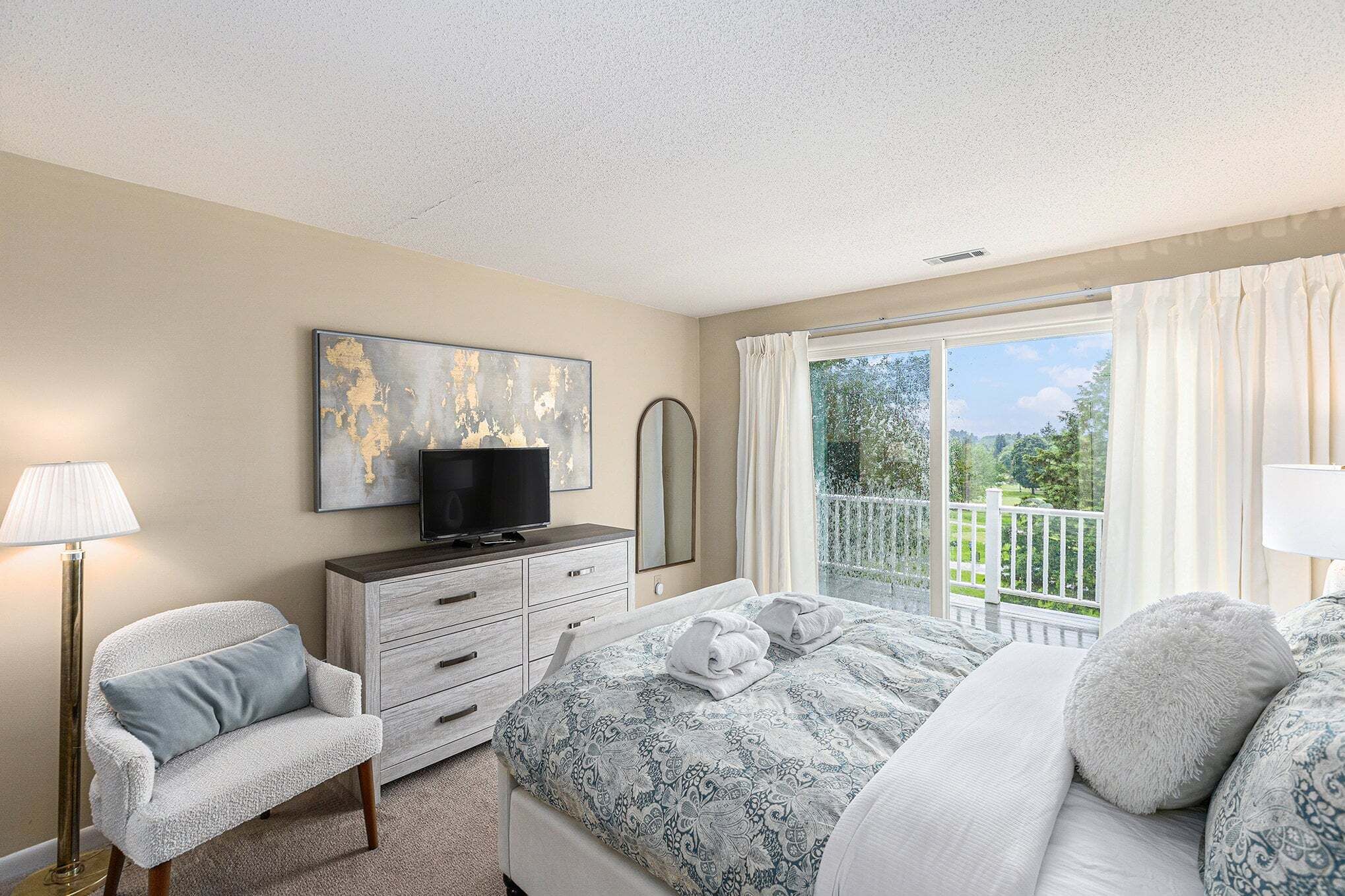 A bedroom with a bed, a dresser, a chair, a TV, and a balcony with a view.