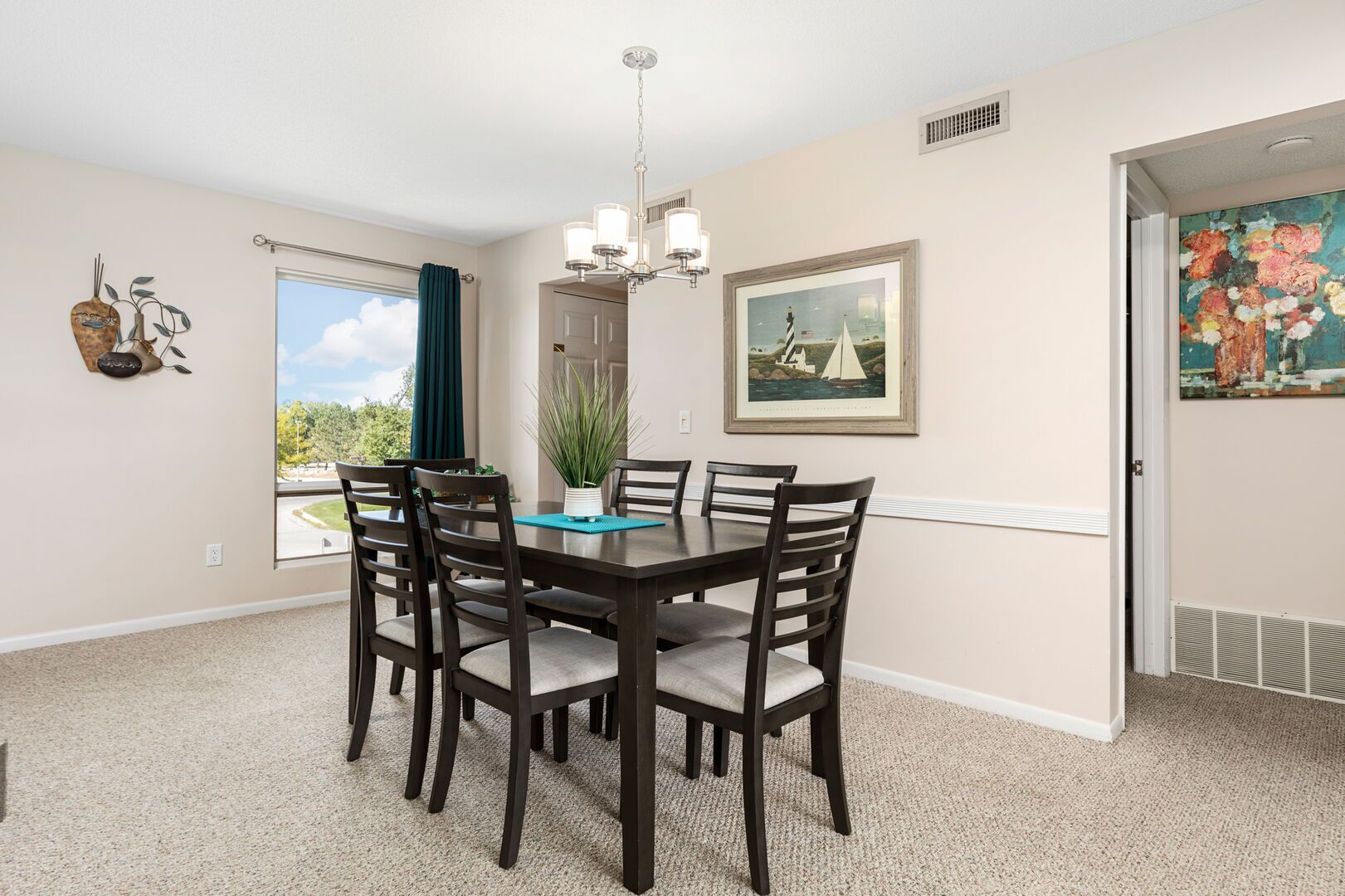 A dining area with a dark wooden table and chairs, a chandelier, a window with a view, and a painting on the wall.
