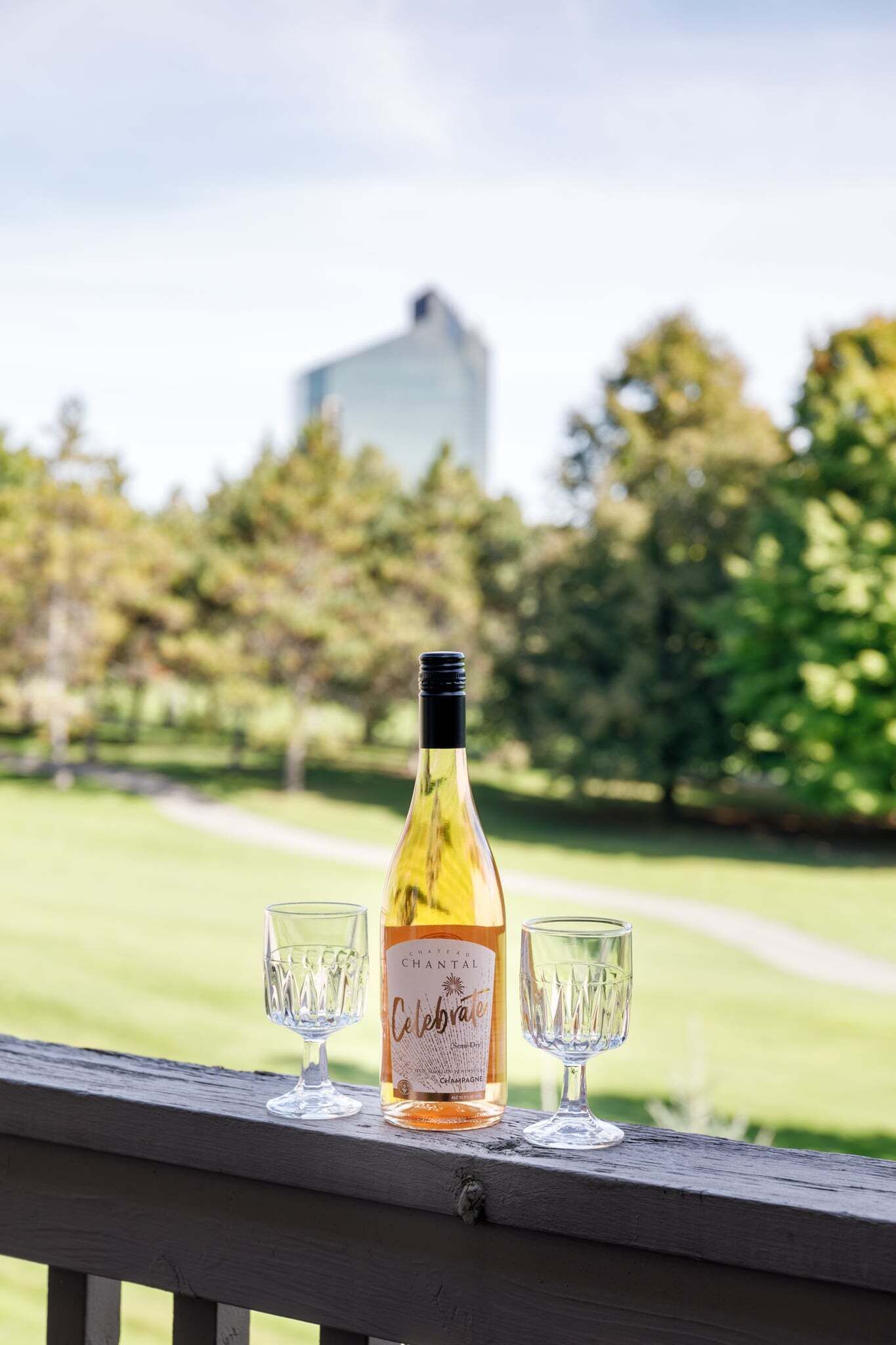 A bottle of Chantal Cuvée wine and two glasses on a wooden railing with a park view in the background.