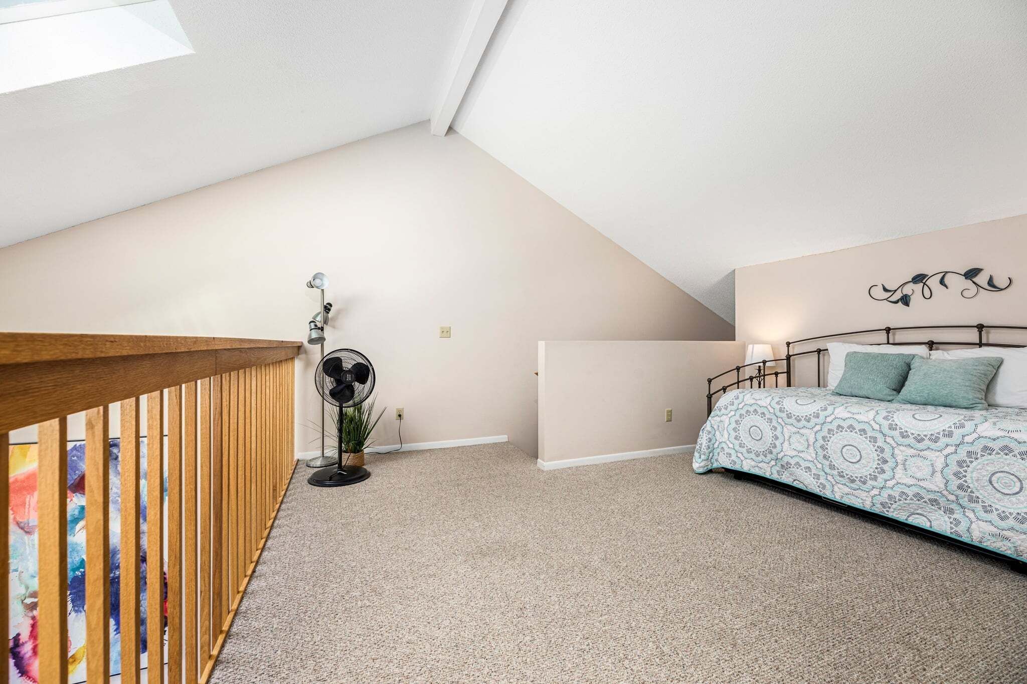 Une chambre avec un plafond en pente, un lit avec une literie à motifs, une rambarde en bois et un ventilateur sur pied.