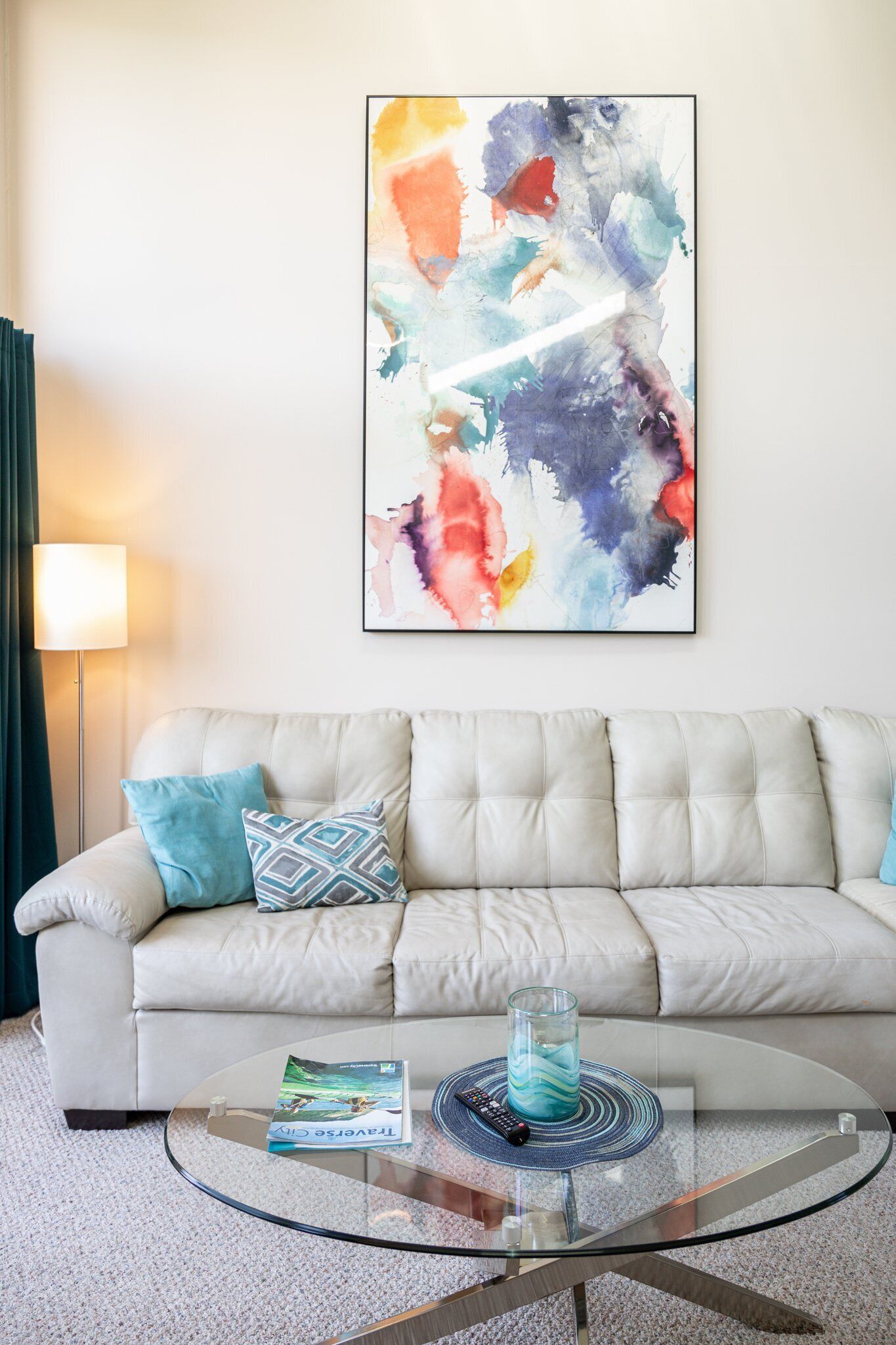 Una sala de estar con un sofá color beige, una mesa de centro de vidrio y un cuadro abstracto de colores en la pared.