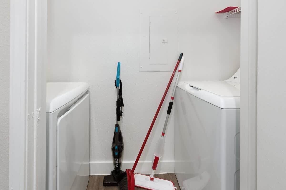 A small, narrow bathroom with a white toilet, a blue and black vacuum cleaner, and a red and white mop leaning against the wall.