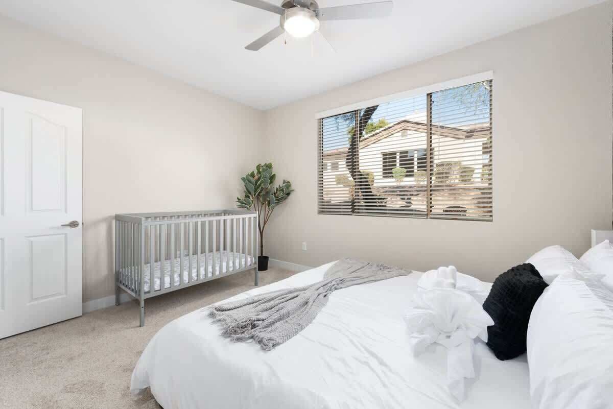 A bedroom with a large bed, a crib, a ceiling fan, and a window with blinds.