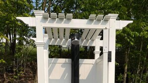 Estructura portátil de baño exterior con una pérgola blanca, un asiento de inodoro negro y un escalón de madera.