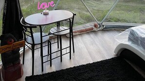 Una habitación con un amplio ventanal de vidrio abovedado, una pequeña mesa redonda con sillas, una cama y un oso de peluche.