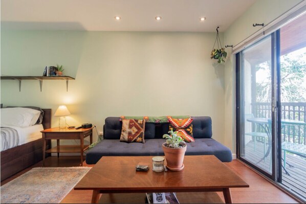 A cozy room with a bed, a sofa, a coffee table, a hanging plant, and a window with a view of the outdoors.