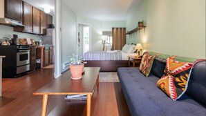 Un espacio habitable compacto con kitchenette, una cama con cabecera, un sofá con cojines estampados y una mesita con planta en maceta.