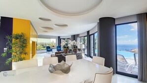 A modern dining area with a white table, blue chairs, and a large window offering a view of the ocean. - Villa Bonsai (Calheta)