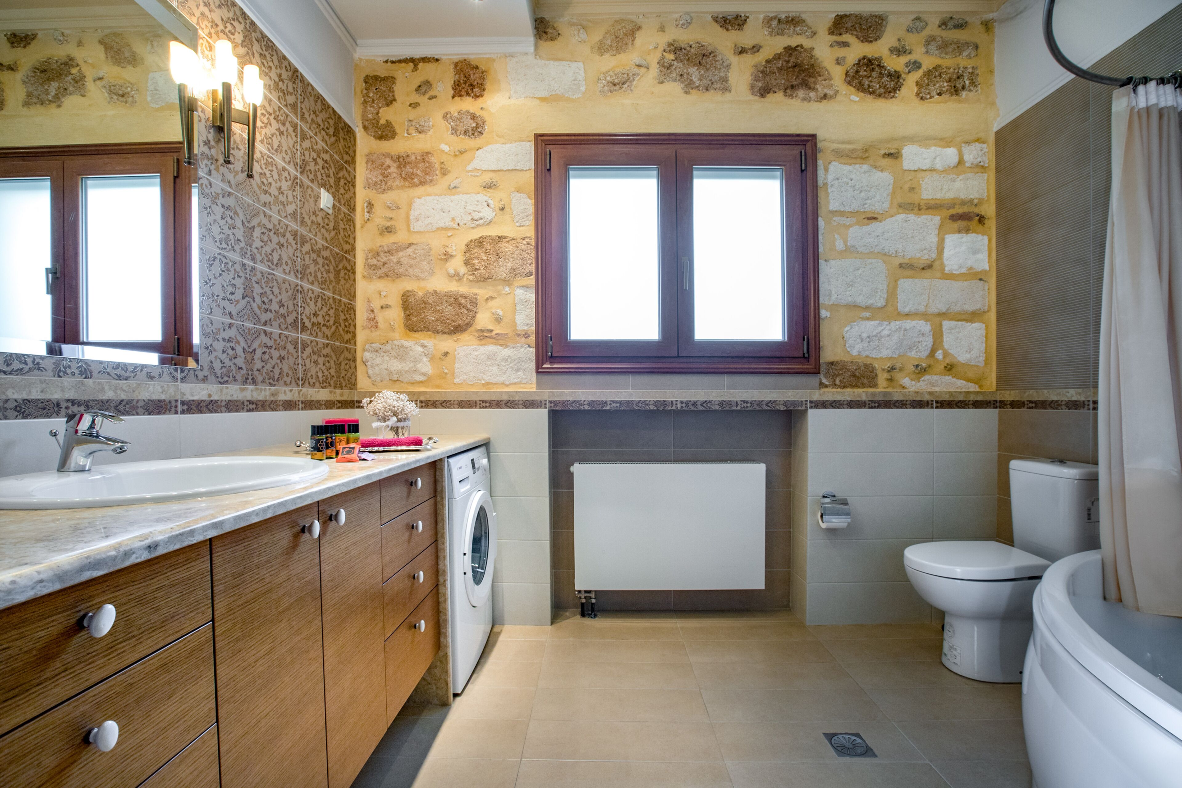 Une salle de bain avec un mur en pierre, un meuble-lavabo en bois, un évier, un lave-linge, des toilettes et une fenêtre avec un rideau.