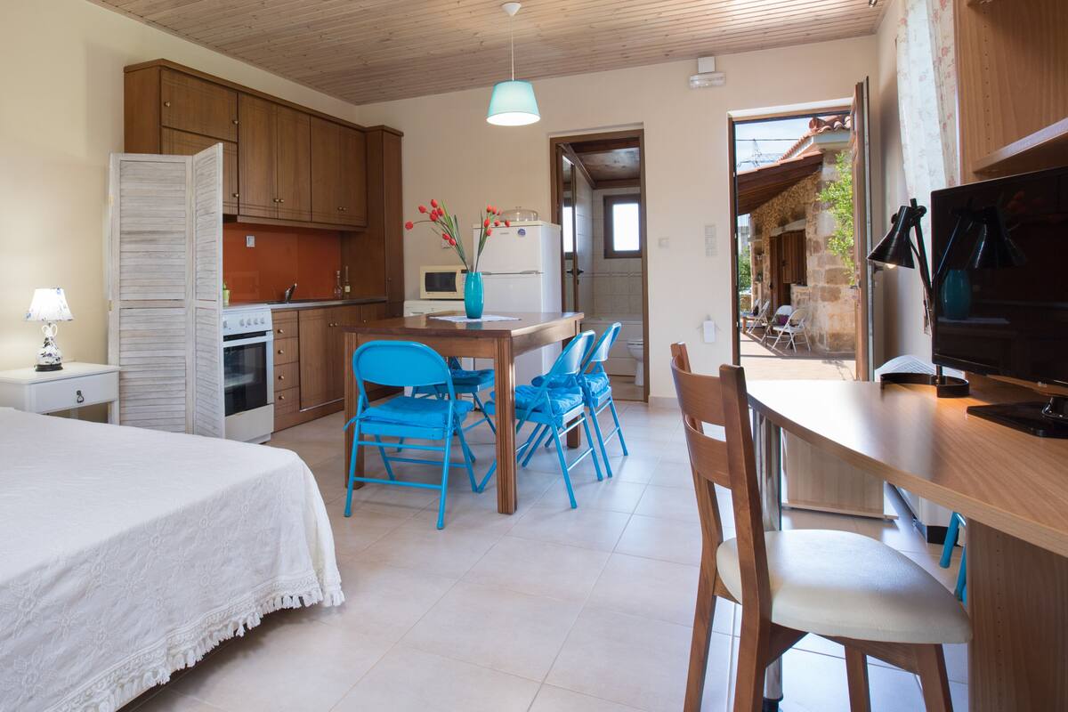 A compact kitchen with wooden cabinets, a dining area with blue chairs, a white bed, and a television.