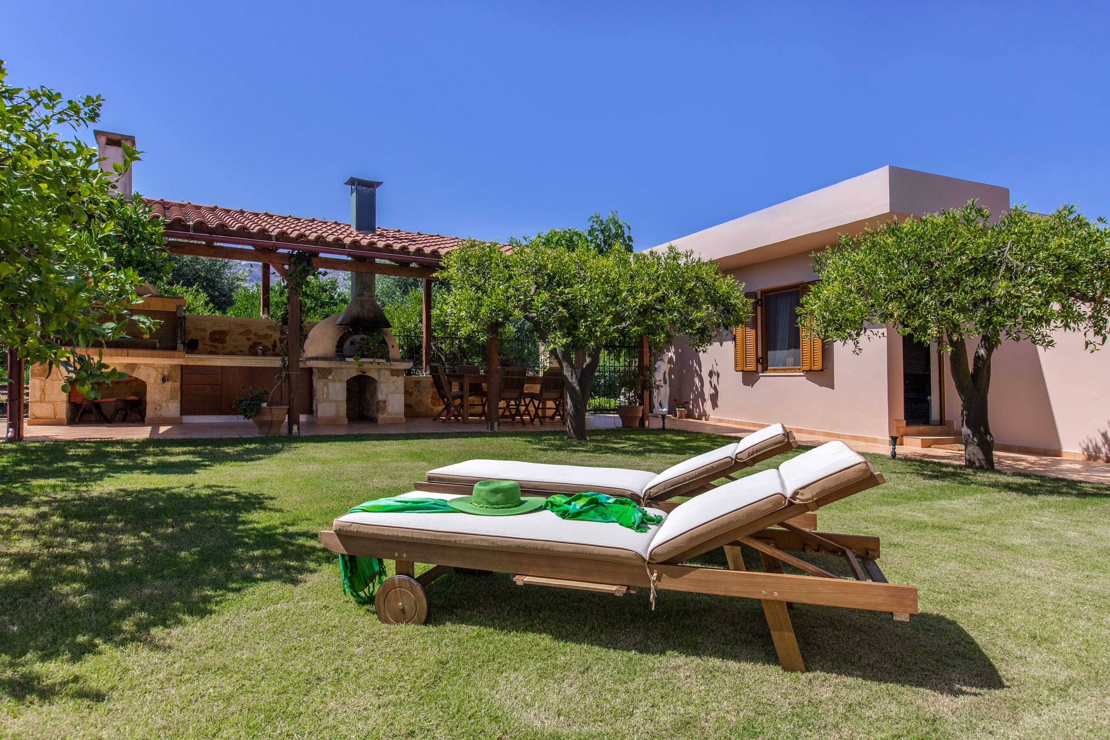 Un patio moderno al aire libre con una silla lounge de madera y una mesa, rodeado de árboles y un edificio con techo de tejas.