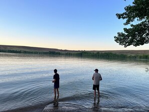 Two people standing in a calm lake with a clear sky and distant hills. - Queen and twin bed Trailer on private beach, Hammocks, tiki torches, fishing (Clifton)