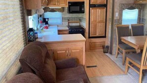 A compact RV kitchen with wooden cabinets, a microwave, stove, and a dining area with a table and chairs. - Queen and twin bed Trailer on private beach, Hammocks, tiki torches, fishing (Clifton)