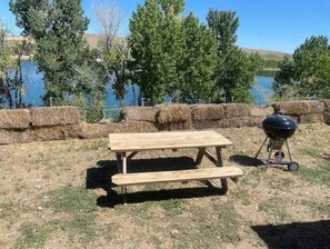 An outdoor picnic setup with a wooden table and benches, a grill, and hay bales in a grassy area with trees and a body of water in the background. - Queen and twin bed Trailer on private beach, Hammocks, tiki torches, fishing (Clifton)