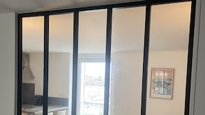 A room with a large window, a kitchen area visible through the window, and a framed picture on the wall.