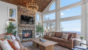 A spacious living room with a stone fireplace, large windows, and a cozy seating area. - Designer Home w/ Incredible Views, Sleeps 16 (Wintergreen Resort)