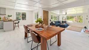 A spacious living area with a wooden dining table, comfortable seating, and a large window offering a view of the outdoors. - Mountain Oasis: Modern Plantation Home (Kealakekua)
