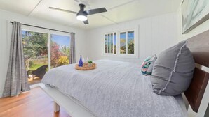 A bedroom with a bed, a ceiling fan, a window with a view of greenery, and a bench. - Mountain Oasis: Modern Plantation Home (Kealakekua)