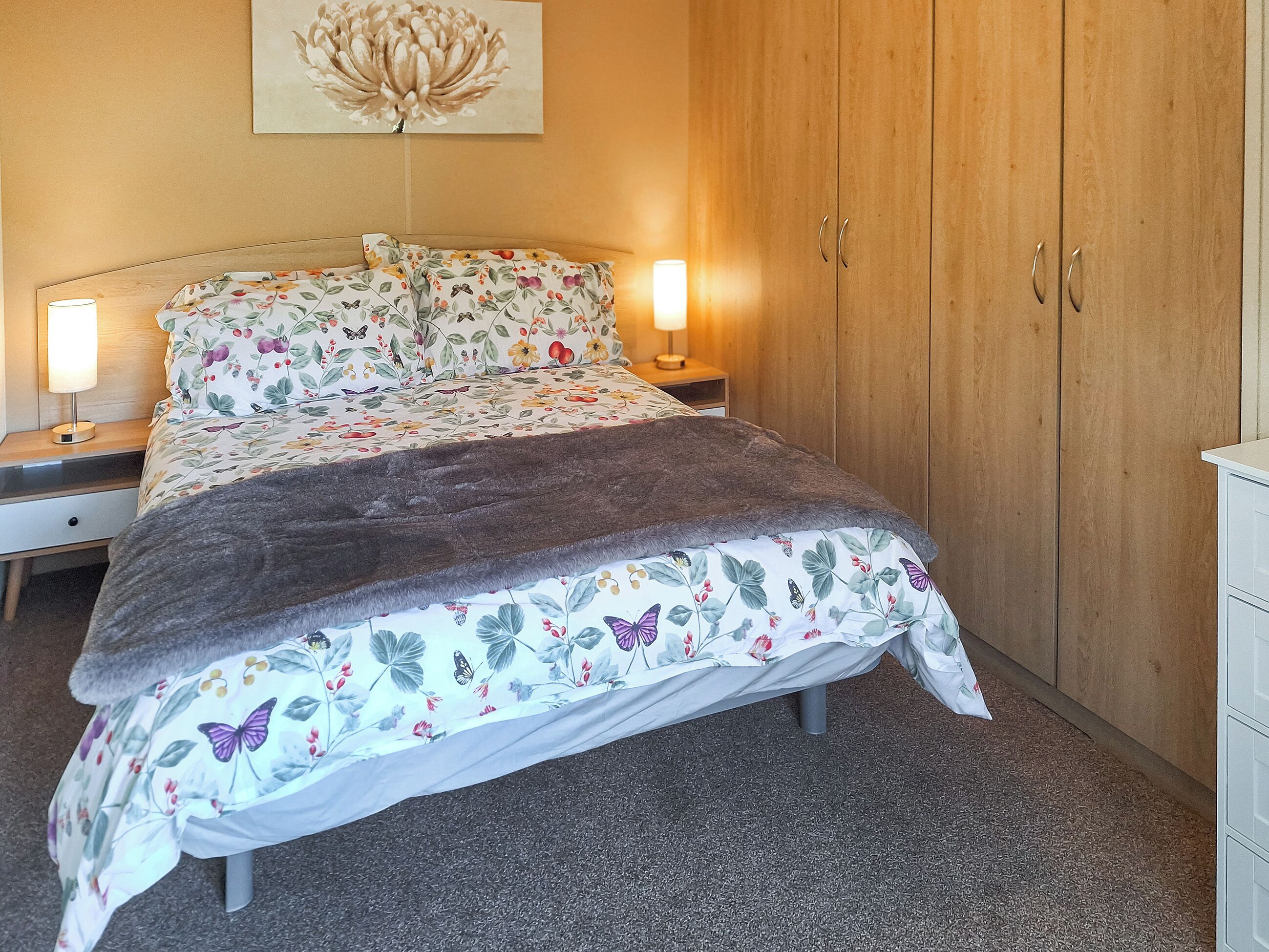 Una cama bien hecha con ropa de cama floral, dos lámparas de noche, un armario de madera y una obra de arte colgada en la pared.
