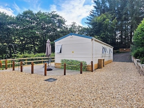 A small, single-story mobile home with a blue roof, surrounded by gravel, wooden fencing, and trees. - 2 bedroom accommodation in New Radnor, near Presteigne (New Radnor, near Presteigne)