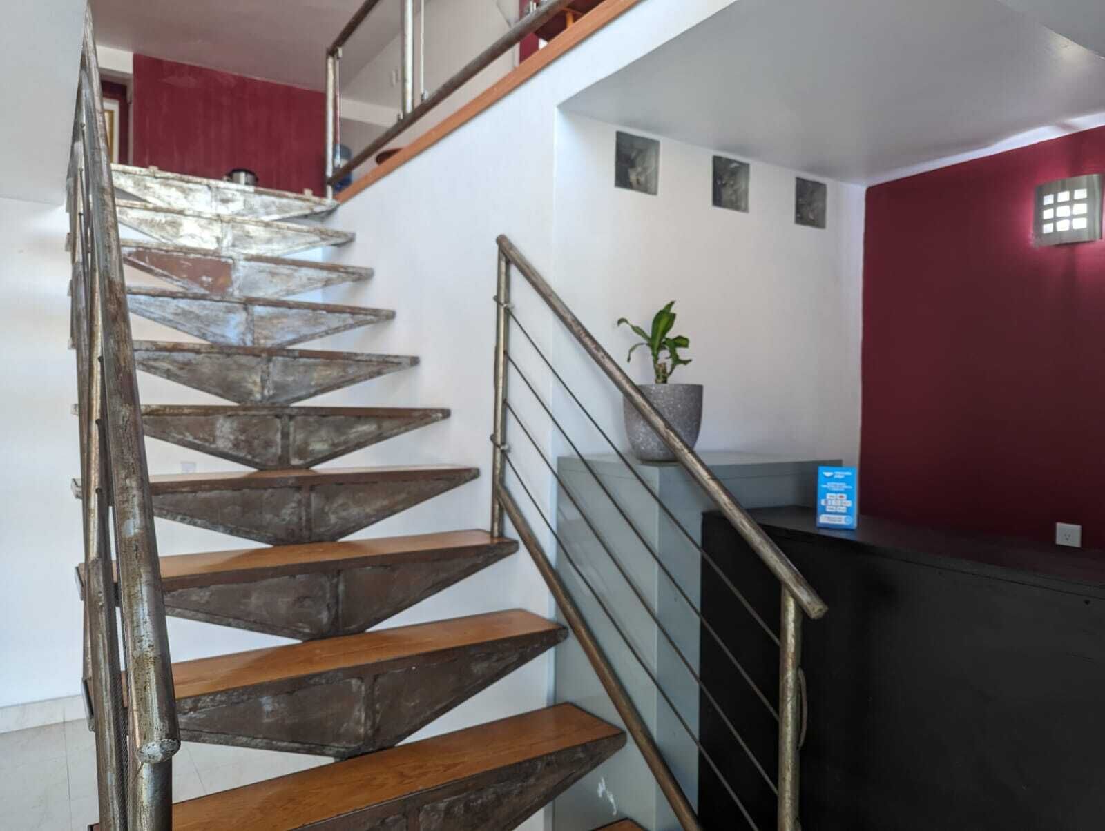 Una escalera de madera con barandales metálicos que conduce a una plataforma con una planta en maceta y un letrero azul.