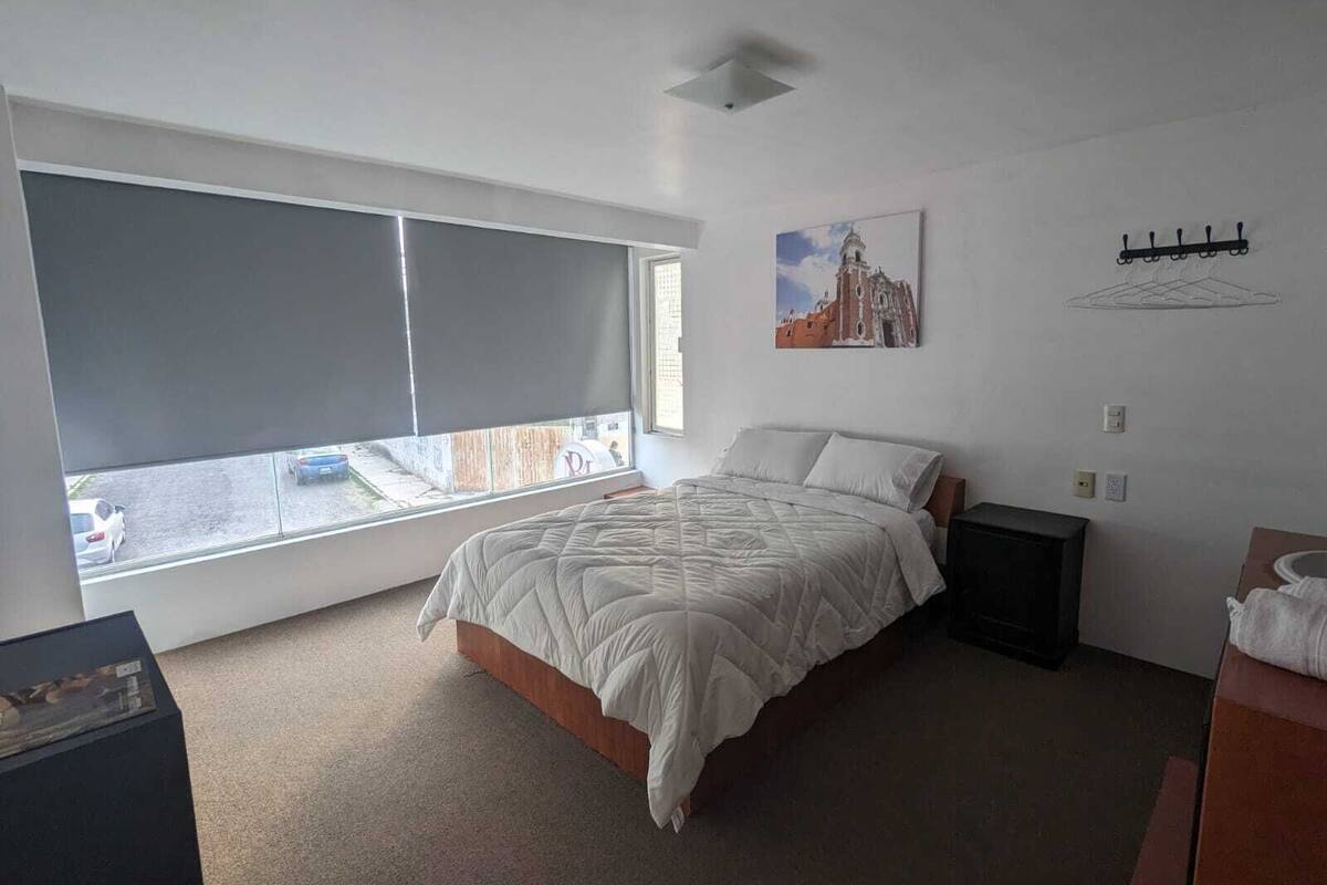 Un dormitorio con un ventanal grande, una cama, una mesita de noche y un cuadro en la pared.