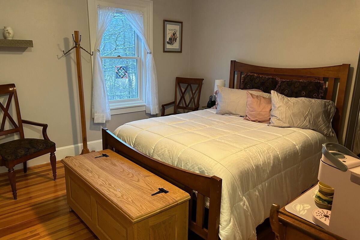 A bedroom with a wooden bed, a wooden chest of drawers, a chair, a bedside table, and a window with curtains.