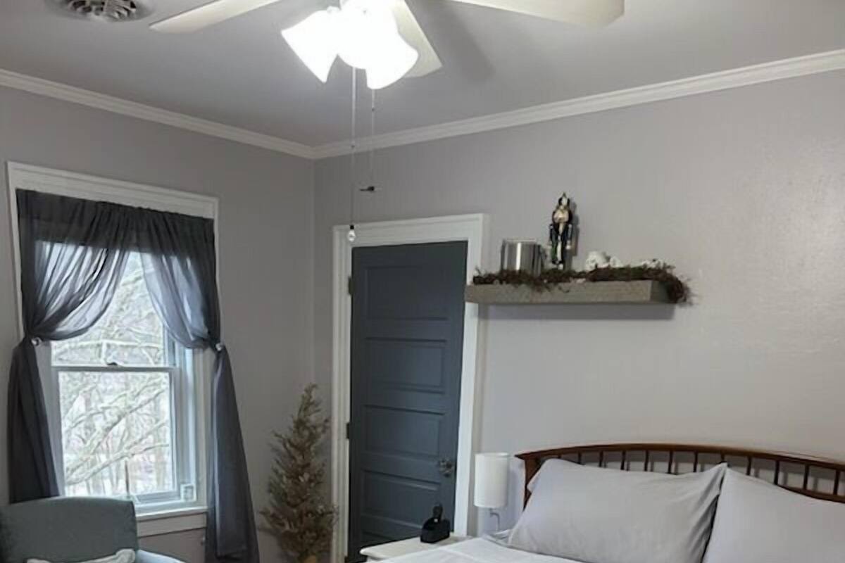 A bedroom with a bed, a chair, a bedside table, a ceiling fan, a window with curtains, and a door.