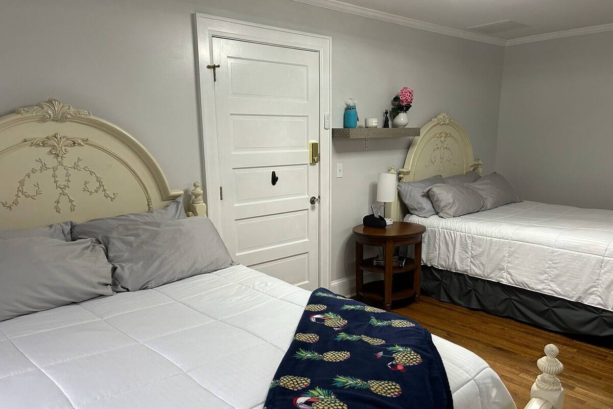 A bedroom with two beds, a white door, a small wooden side table, and a shelf with decorative items.