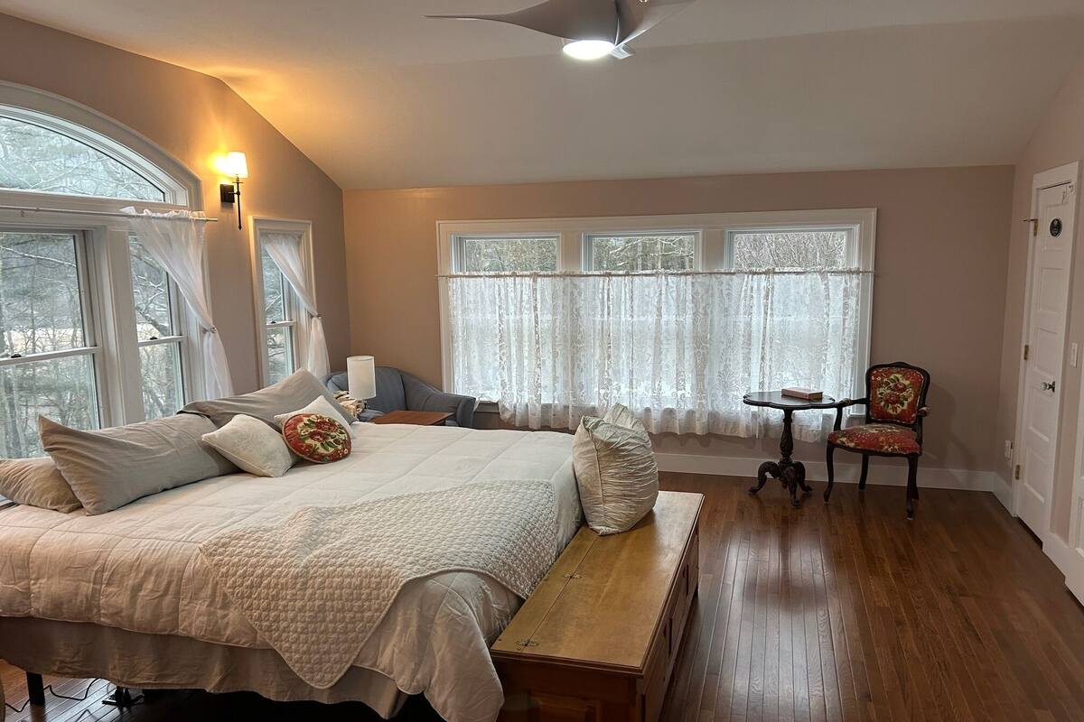 A bedroom with a large bed, a wooden bench, a desk, and a chair. There are windows with curtains, a ceiling fan, and a door.