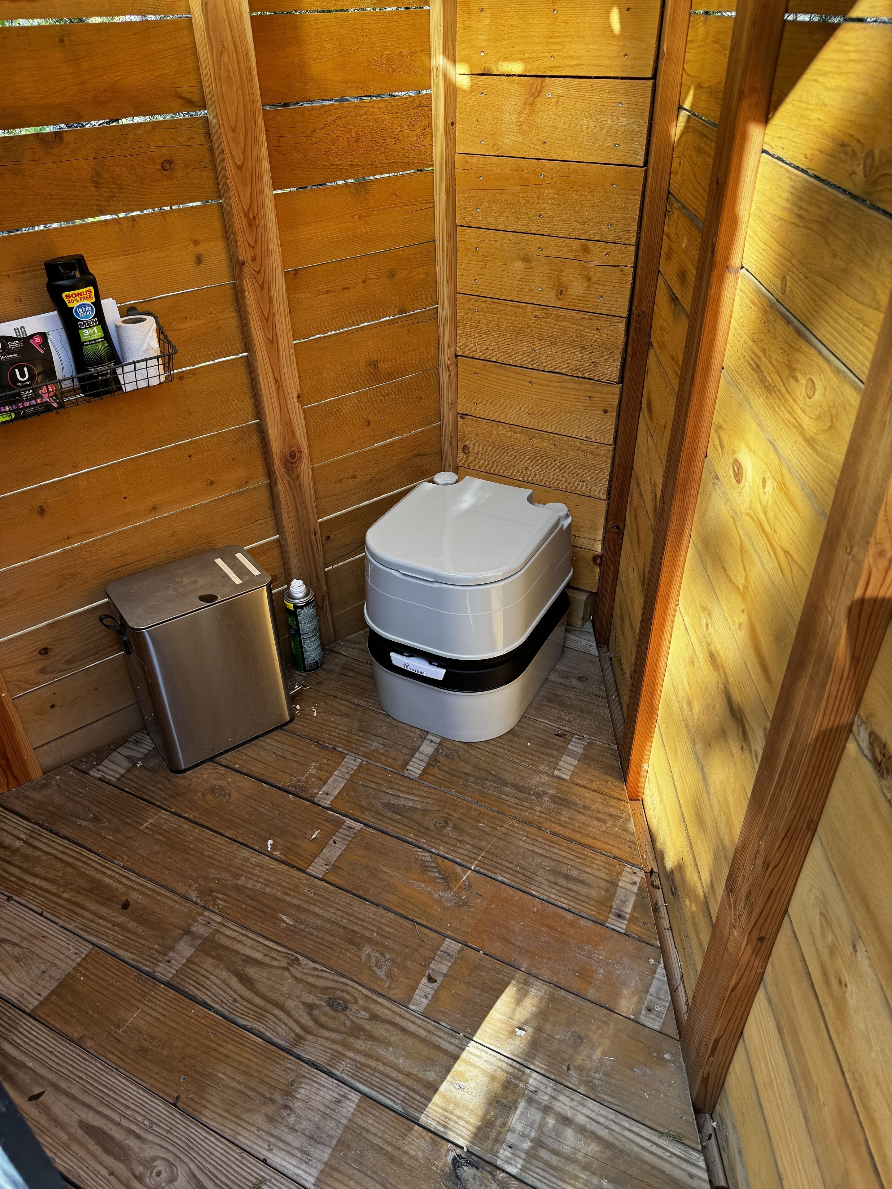 A compact wooden bathroom with a toilet, a small trash bin, and a shelf with toiletries.
