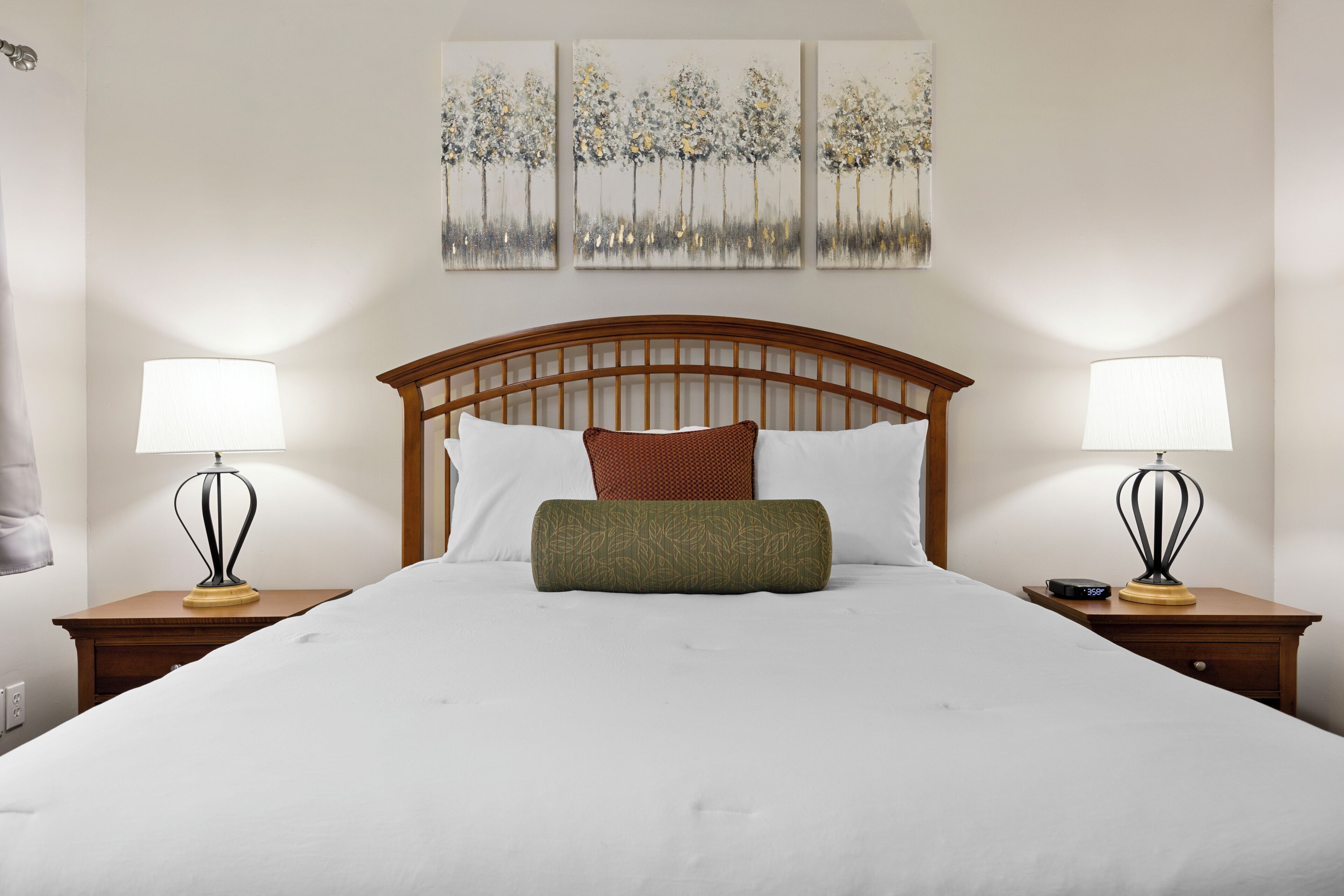 A neatly made bed with a wooden headboard, two bedside tables with lamps, and three wall-mounted paintings.