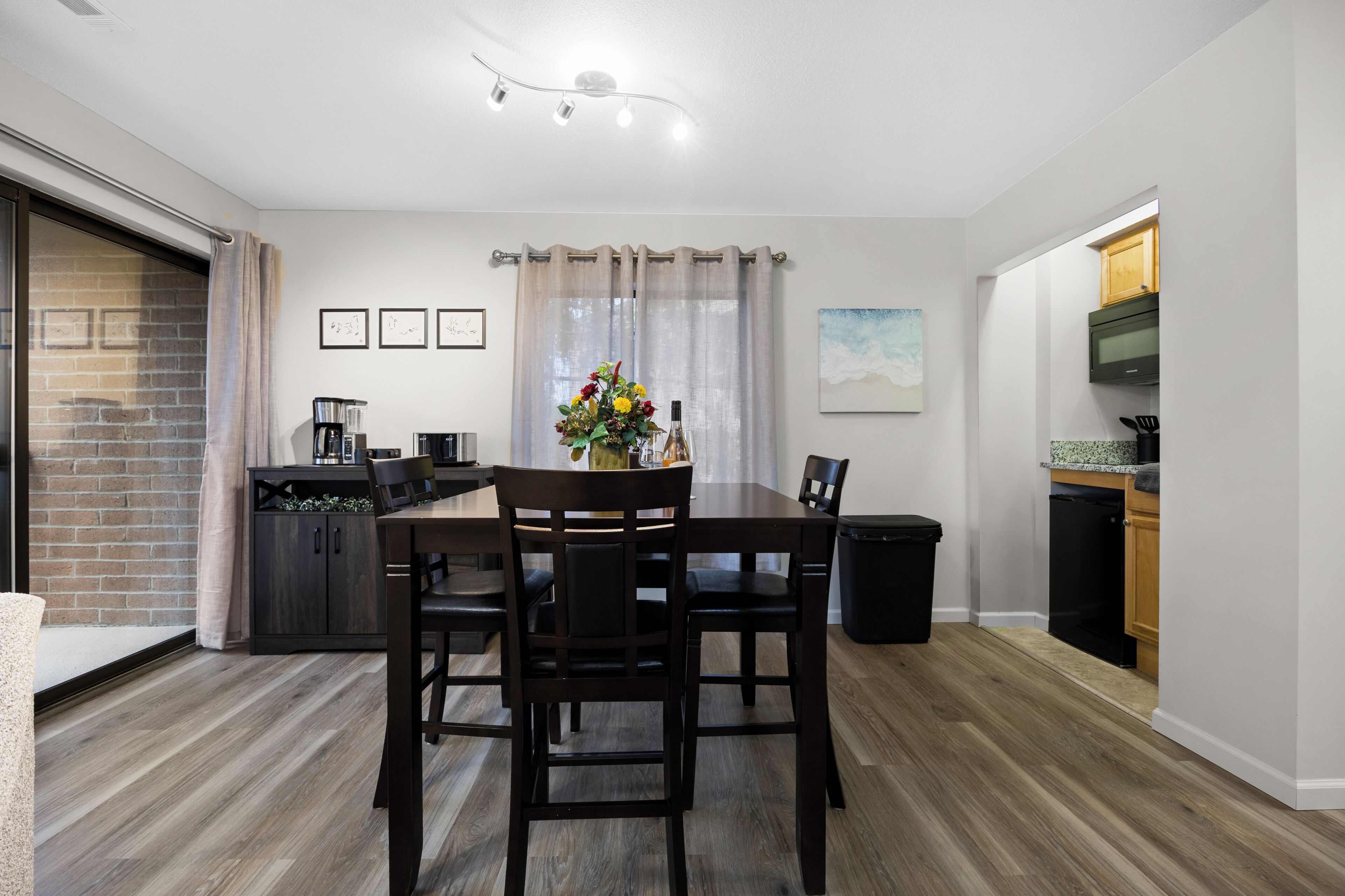 A dining area with a wooden table, chairs, a vase with flowers, a coffee machine, and a TV mounted on the wall.
