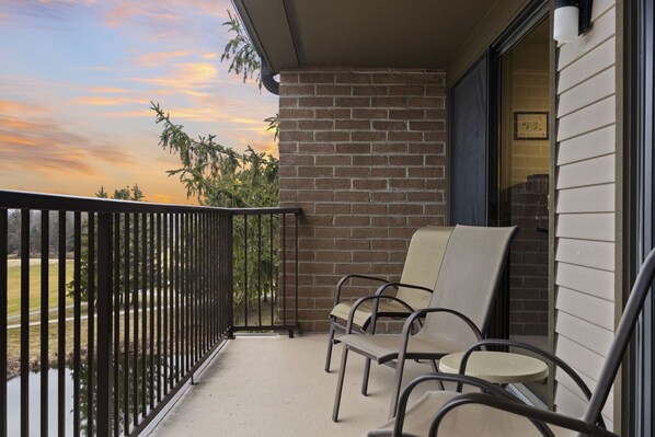 A balcony with a brick wall, two chairs, and a view of a park and sunset. - Lakeview Golf Retreat 1BDR Condo 5057 (Williamsburg)