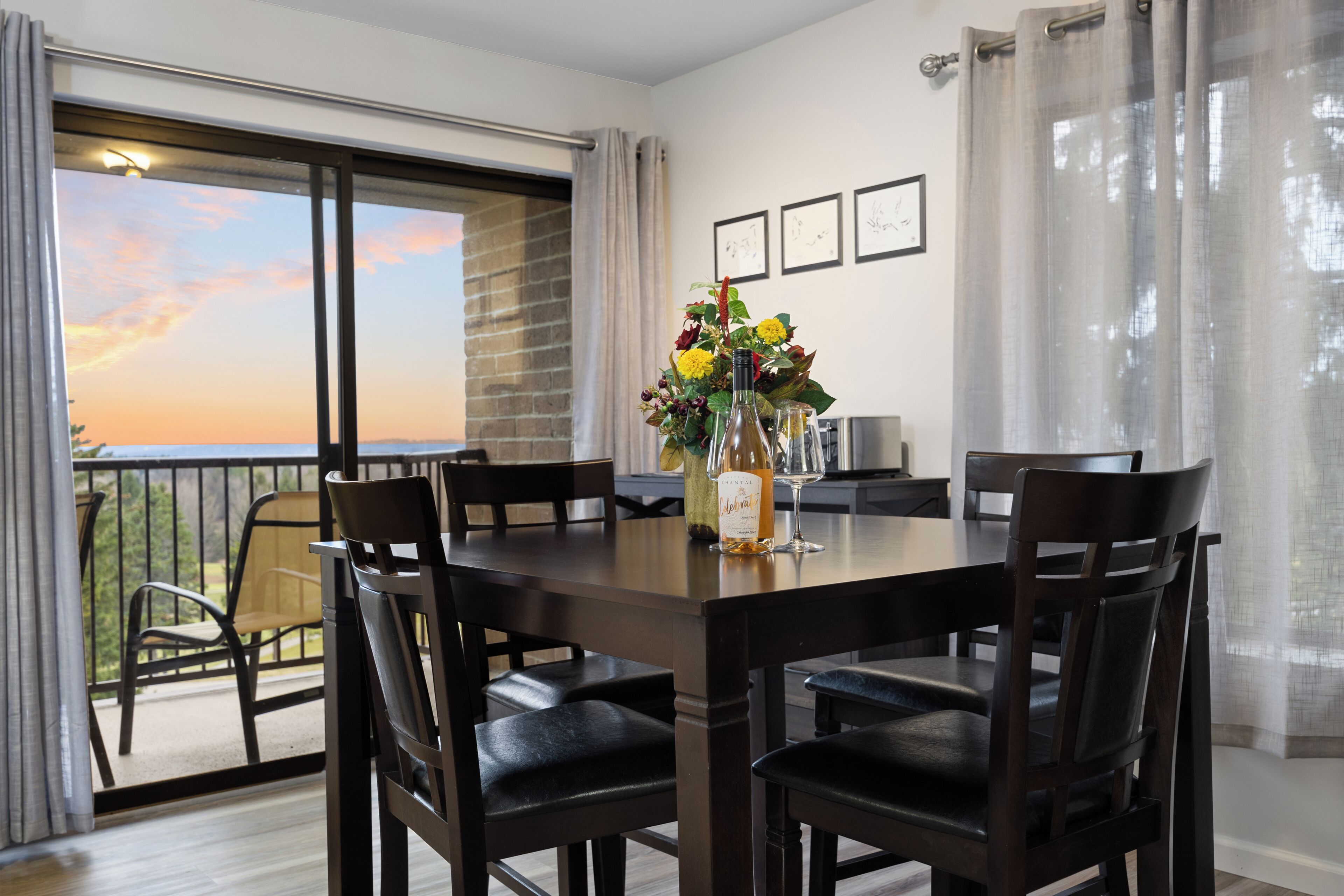 Un comedor con una mesa y sillas, un jarrón con flores y vistas al atardecer desde una puerta balcón.