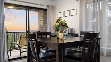 Un comedor con una mesa y sillas, un jarrón con flores y vistas al atardecer desde una puerta balcón.