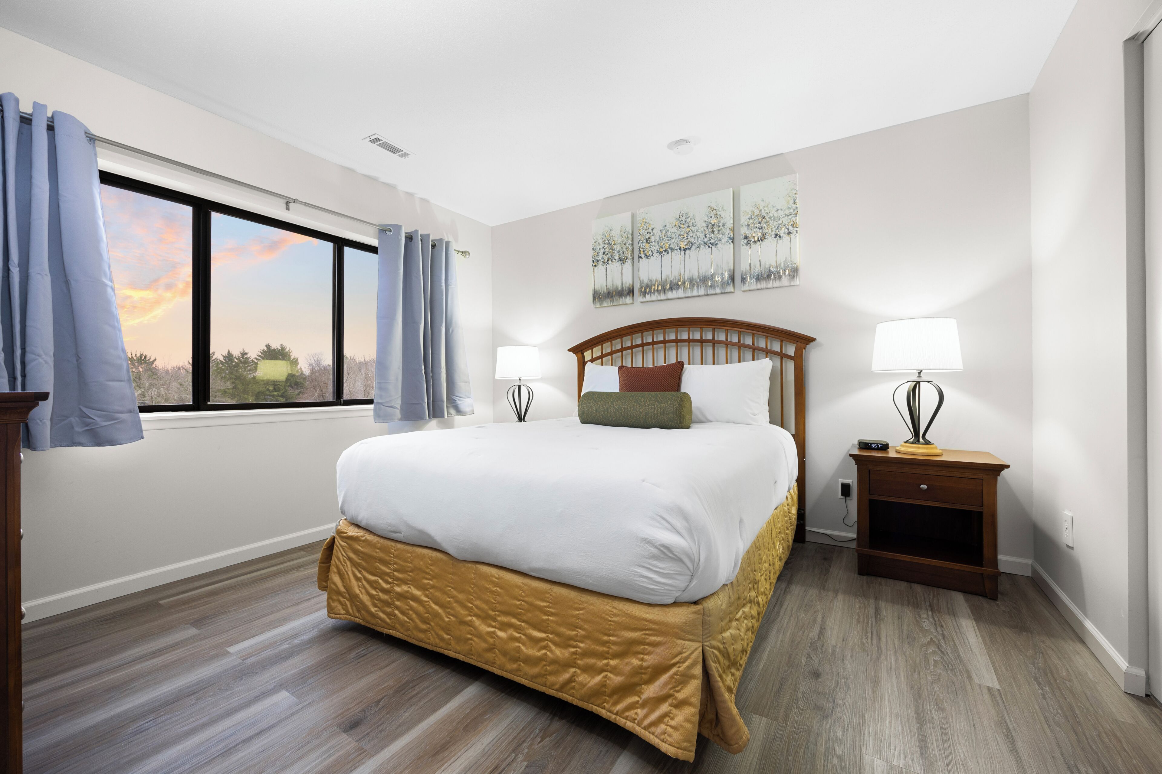A hotel room with a large bed, two bedside tables, a window with curtains, and a painting on the wall.