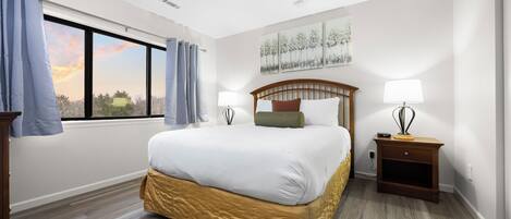 A hotel room with a large bed, two bedside tables, a window with curtains, and a painting on the wall.
