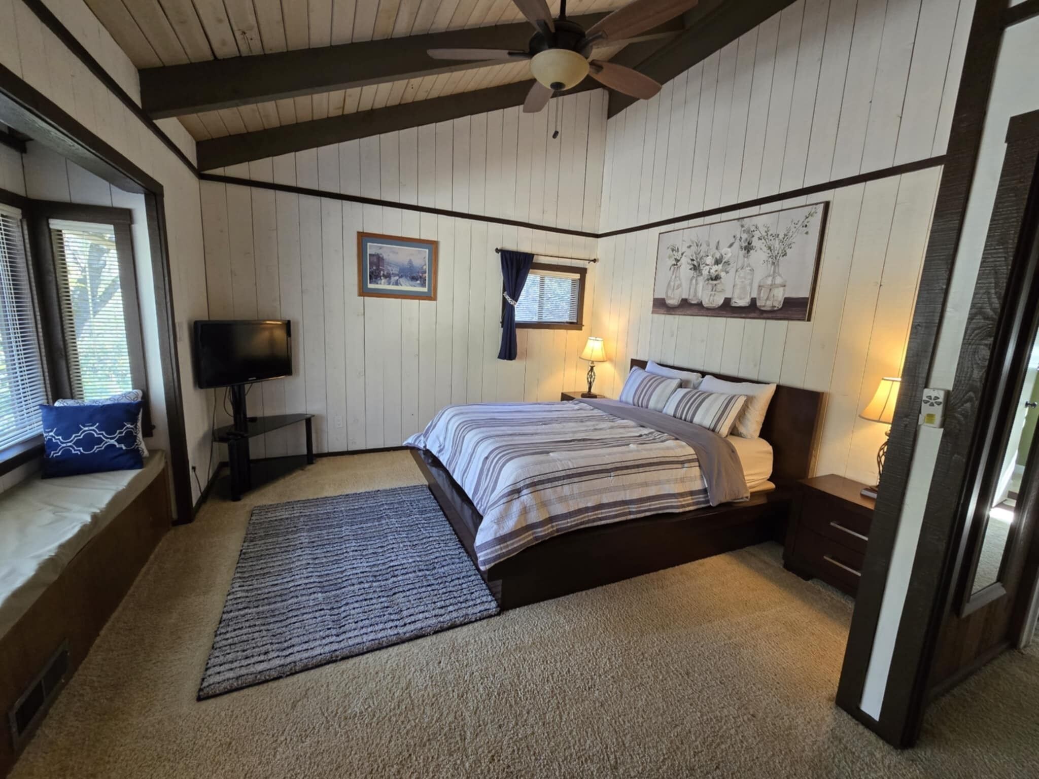 A bedroom with a bed, a television, a ceiling fan, and a window with blinds.