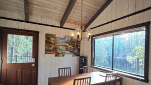 Un comedor con una mesa y sillas de madera, una araña, un cuadro y ventanas amplias con vista a árboles.