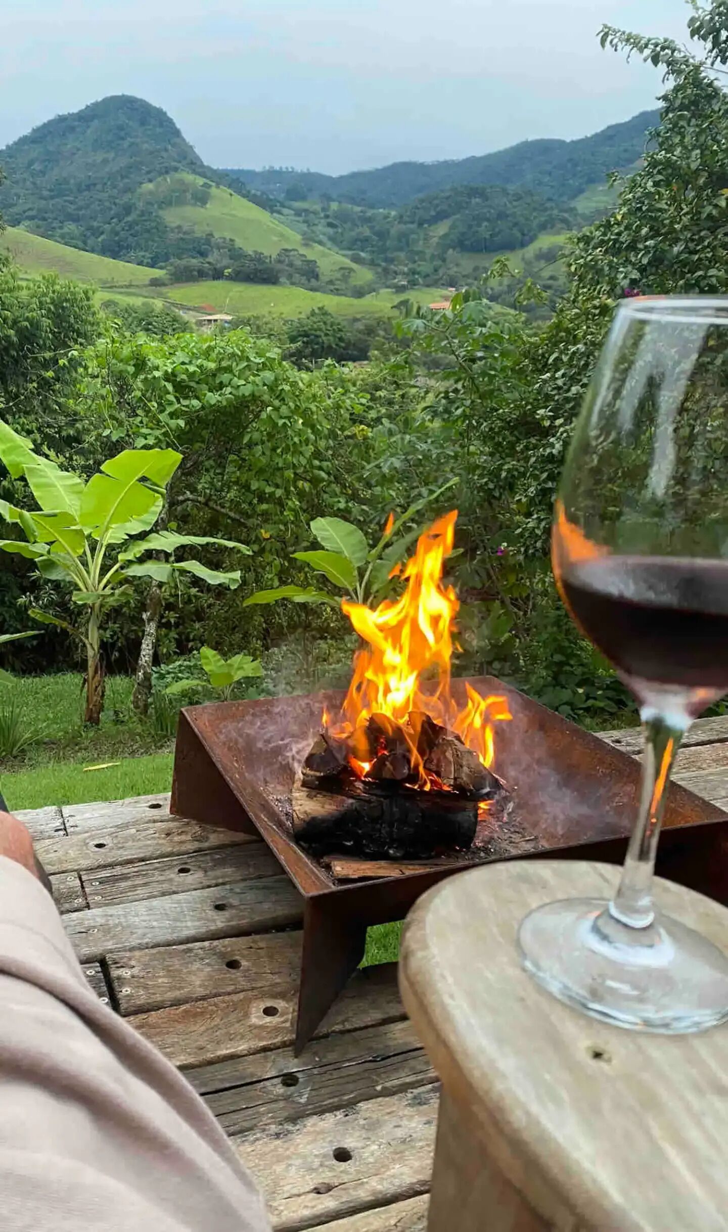 A wooden deck with a metal fire pit and a glass of red wine, overlooking a lush green landscape with hills.
