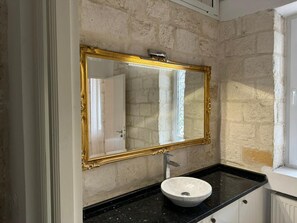Un baño con pared de piedra, espejo con marco dorado, lavabo blanco y encimera negra.